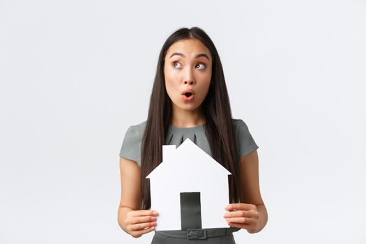 Insurance, loan, real estate and family concept. Surprised and amazed asian female holding paper house and looking upper left corner amazed, searching perfect home, white background.