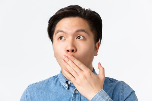 Close-up of shocked and startled young asian guy gasping of amazement, looking upper left corner, cover opened mouth in awe, standing impressed over white background.