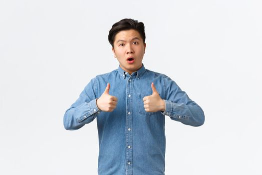 Impressed and astounded handsome asian guy showing thumbs-up and looking astonished at camera, congrats person with excellent work, unexpected good work, saying well done, white background.