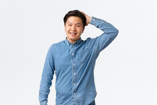 Modest smiling asian man, blushing as making good deed, scratching head confused and awkward, standing cute over white background. Nice guy talking to pretty lady. Copy space