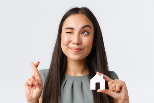 Insurance, loan, real estate and family concept. Close-up of hopeful smiling asian woman looking upper left corner, fingers crossed and making wish, showing small paper house, white background.