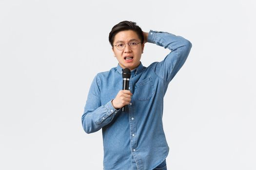 Lifestyle, leisure and people concept. Shy and insecure asian guy standing indecisive over white background with microphone, scared of giving speech in public, standing anxious white background.