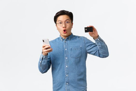 Impressed and excited asian bank customer, guy in glasses and casual clothes showing credit card while using smartphone to open e-banking application, shopping in internet with mobile app.