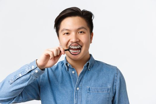 Orthodontics, dental care and stomatology concept. Happy handsome asian young man smiling, open mouth and showing teeth braces with magnifying glass, standing white background.