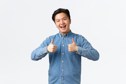 Satisfied happy asian man with braces, showing thumbs-up in approval, like and recommend product or service, congratulating friend with achievement, saying well done, excellent, white background.