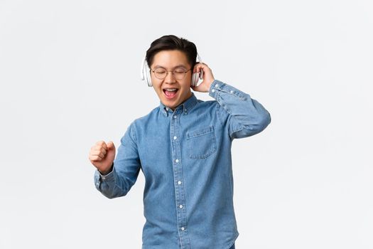 Lifestyle, leisure and technology concept. Happy dancing asian man in glasses, listening music wireless headphones, singing along, having fun with new album on streaming platform.