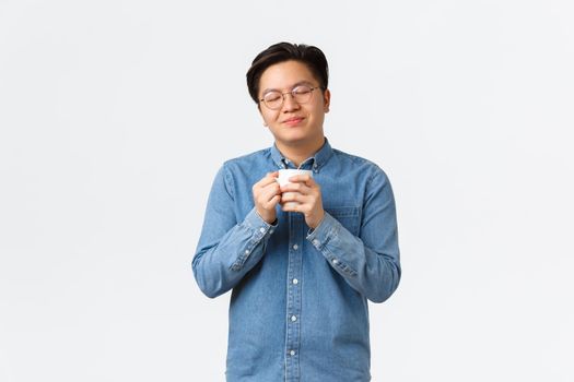 Relaxed and happy smiling asian man with closed eyes enjoying delicious aroma of coffee, holding mug and drinking cappuccino, morning routine and lifestyle concept.