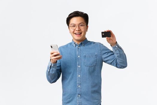 Smiling happy asian guy in glasses and casual clothes using mobile phone to transer money to bank account, showing credit card with satisfied expression, standing pleased white background.