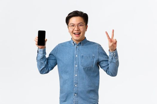 Smiling joyful asian male freelancer, student in glasses and braces, showing smartphone screen and peace sign, standing satisfied with new appllication. Man recommends download app.