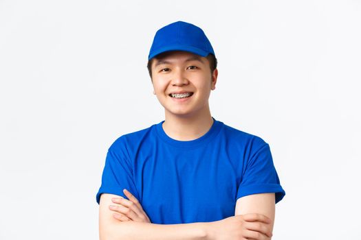 Online shopping, fast shipping, employees and home delivery concept. Upbeat confident asian male courier in blue uniform cap, t-shirt, cross amrs chest and smiling, white background.