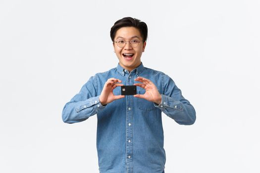 Excited smiling asian guy introduce new banking feature, recommend service, standing in glasses and braces, showing credit card, ready for shopping, paying contactless, white background.