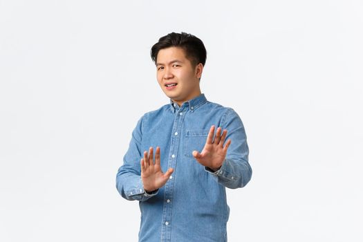 No thank you. Unamused and bothered asian man step away from trouble, raising hands in stop gesture, refusing or rejecting ofer, denying something bad, standing white background.