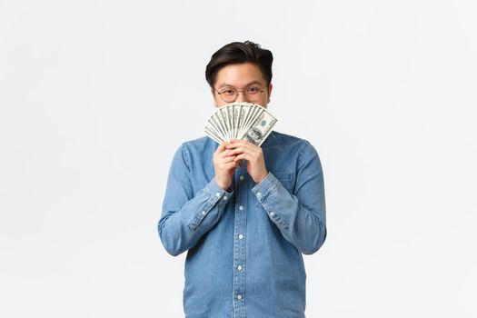 Smiling satisfied asian guy in glasses, hiding face behind money and peeking at camera cunning, earn cash for working freelance, making investment, close deposit, standing white background.