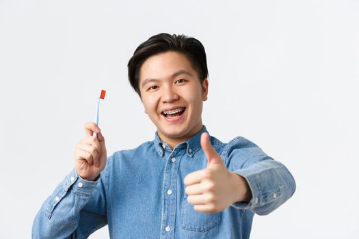 Orthodontics, dental care and hygiene concept. Close-up of satisfied happy asian guy holding toothbrush, smiling with teeth braces and showing thumbs-up in approval, white background.
