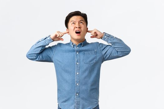 Annoyed and bothered asian man complaining on loud neighbours, shut ears with fingers, looking up displeased, shouting at people upstairs, asking turn-off annoying music, standing white background.