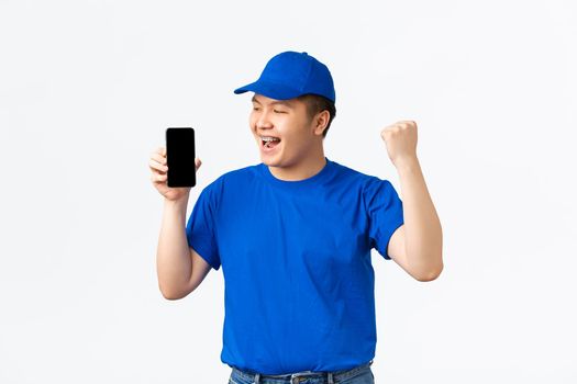 Technology, online shopping and shipping concept. Triumphing cheerful asian delivery man in blue courier uniform, showing fist pump and looking at mobile phone screen, celebrating.