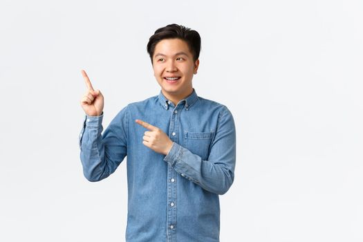 Smiling young asian cute guy in shirt pointing and looking upper left corner pleased face, making announcement, give advice. Man making choice at store, recommend promo banner, white background.