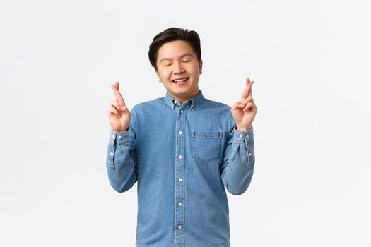 Smiling hopeful asian man feeling lucky, close eyse and grinning as awaiting for dream come true. Guy with braces cross fingers good luck, praying, asking god favour, standing white background.