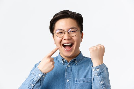 Orthodontics and stomatology concept. Close-up of satisfied happy asian man pointing at his dental braces and smiling broadly, fist pump, rejoicing, fixing teeth, standing white background.