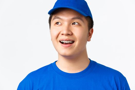 Online shopping, fast shipping, employees and home delivery concept. Close-up of dreamy, happy smiling asian male courier in blue uniform, turn head left and looking thoughtful, white background.
