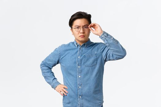 Serious-looking suspicious asian male entrepreneur touching glasses on face and frowning doubtful, standing hesitant or skeptical, dont trust someone, posing uncertain white background.