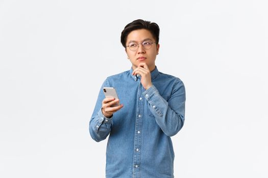 Thoughtful creative asian man in glasses thinking while making post on social media, looking away, pondering or making decision, holding smartphone, choosing something in internet.