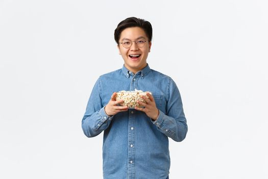 Leisure, lifestyle and people concept. Smiling cute asian man prepared bowl of popcorn for movie night, standing upbeat and grinning, watching TV premier, standing white background.