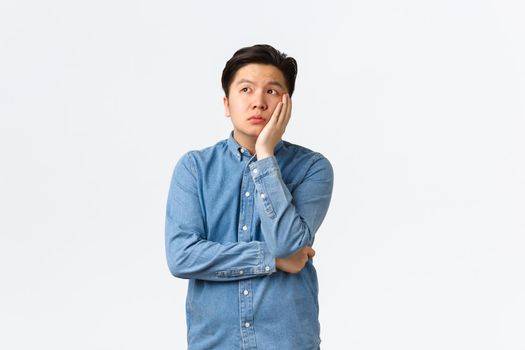 Bored and exhausted asian male student feeling unamused, space out at lecture, leaning face on hand and looking away dreamy, feeling boredom, standing white background, attend boring class.