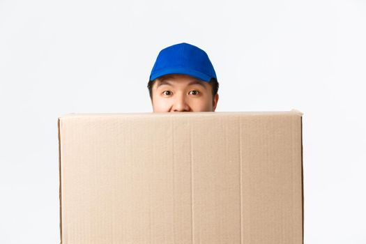 Online shopping, fast shipping concept. Close-up of funny asian delivery man in blue cap appear above large box, carry parcels to client doorstep, handing over package, smiling cute.
