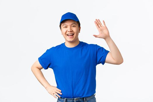 Friendly-looking asian male courier in blue uniform smiling and waiving hand to say hello, greeting customer bring purchase to client doorstep. Cheerful delivery guy make hi gesture, white background.
