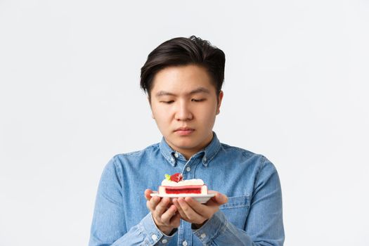 Close-up of gloomy asian guy tempting to try piece cake, looking at dessert with desire. Man on diet trying resist temptation of eating carbs, losing weight, standing white background.