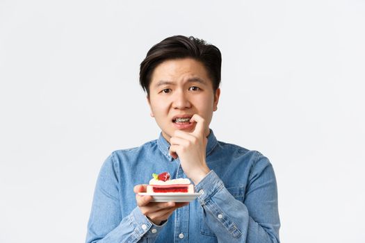 Close-up of tensed and worried asian guy tempting to eat piece of cake while on diet, biting finger indecisive. Man with braces wants bite of dessert, standing white background gloomy.
