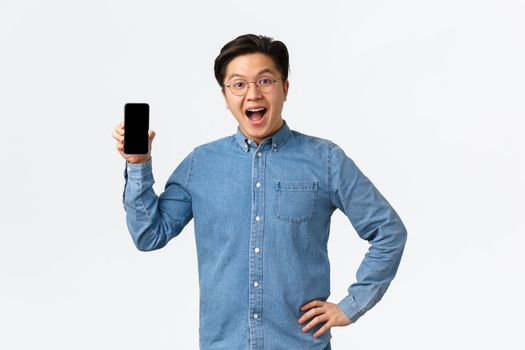 Happy and amused handsome asian guy with braces and glasses react to fantastic news, showing mobile phone screen, introduce application or shop, standing white background amazed.