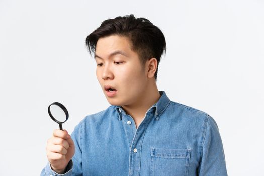 Close-up of young asian guy looking curious and focused, searching for something, looking through magnifying glass down, open mouth intrigued, standing white background. Copy space