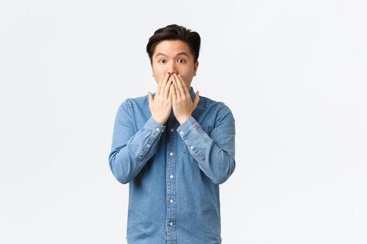 Shocked astounded asian man witness accident, gasping and cover mouth, gossiping with coworkers at office, hear stunning news, standing white background speechless.