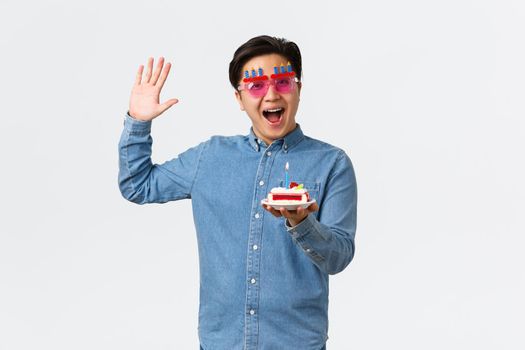 Celebration, holidays and lifestyle concept. Happy smiling friend in funny party sunglasses bring birthday cake with lit candle, enjoying b-day, having fun, standing white background.