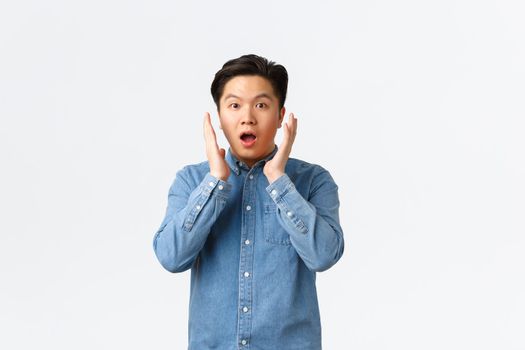 Surprised and impressed asian guy react to big announcement, holding hands near face and gasping, stare at something unbelievable, standing astounded over white background.