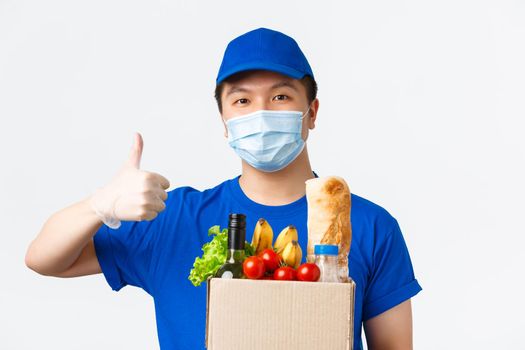 Online shopping, food delivery and covid-19 pandemic concept. Smiling confident asian male courier in medical mask and gloves, blue uniform, carry order, show thumb-up, handing box with groceries.