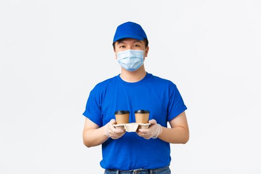 Contactless delivery, covid-19 shipping, preventing virus concept. Friendly smiling young asian male courier in medical mask, gloves and blue uniform, holding takeaway coffee order, white background.