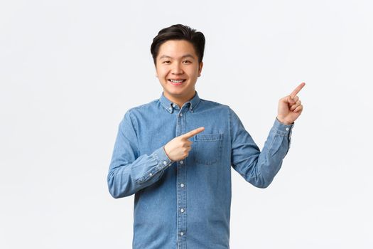 Cheerful smiling asian young man with braces in blue shirt, pointing fingers upper right corner proud and confident, showing link or make announcement, standing white background.