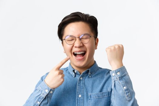 Orthodontics and stomatology concept. Close-up of satisfied happy asian man pointing at his dental braces and smiling broadly, fist pump, rejoicing, fixing teeth, standing white background.