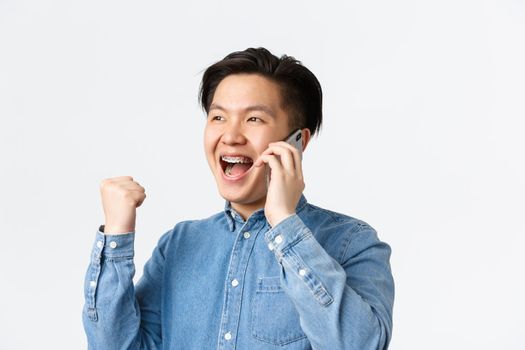 Close-up of winning successful asian businessman in shirt, rejoicing while having conversation over mobile phone, fist pump and smiling, receive good news, standing white background.