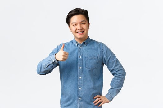 Smiling pleased asian male student with braces, showing thumbs-up, recommend product or service with excellent quality, like and approve idea. Man nod in approval, agree with person, white background.