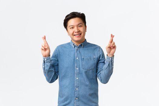 Smiling cute asian man having faith, believe dreams come true, cross fingers good luck, praying or making wish, standing lucky over white background, anticipating positive results.