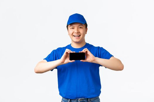 Technology, online shopping and shipping concept. Smiling friendly asian delivery man in blue uniform, standing over white background and showing smartphone screen, mobile application.