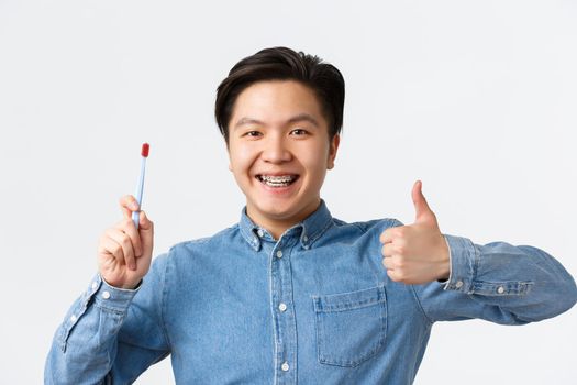 Orthodontics, dental care and hygiene concept. Close-up of satisfied asian man showing thumbs-up while recommend using toothbrush or toothbaste for teeth with braces, smiling pleased.