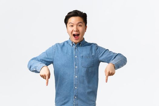 Amazed happy asian guy making huge announcement, telling big awesome news, pointing fingers down at banner. Man showing advertisement with pleased expression, recommend company.