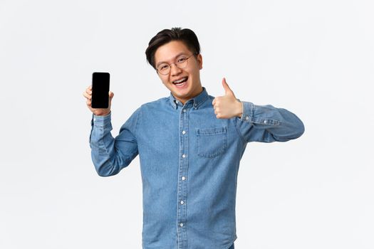Upbeat and satisfied asian man with braces, wearing glasses and casual clothes, showing thumb-up in approval of smartphone application, facing mobile phone screen to promote app, white background.