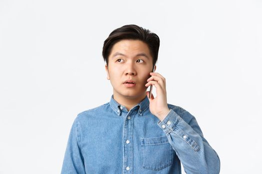 Close-up of indecisive asian guy spacing out during phone call, having conversation and looking away confused, standing white background. Man holding smartphone near ear, order food delivery.