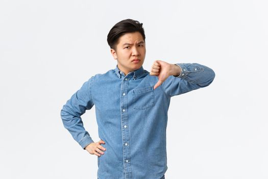 Disappointed skeptical asian man looking judgemental and unamused, standing white background shaking head and showing thumbs-down in disapproval, dislike and dont recommend something.